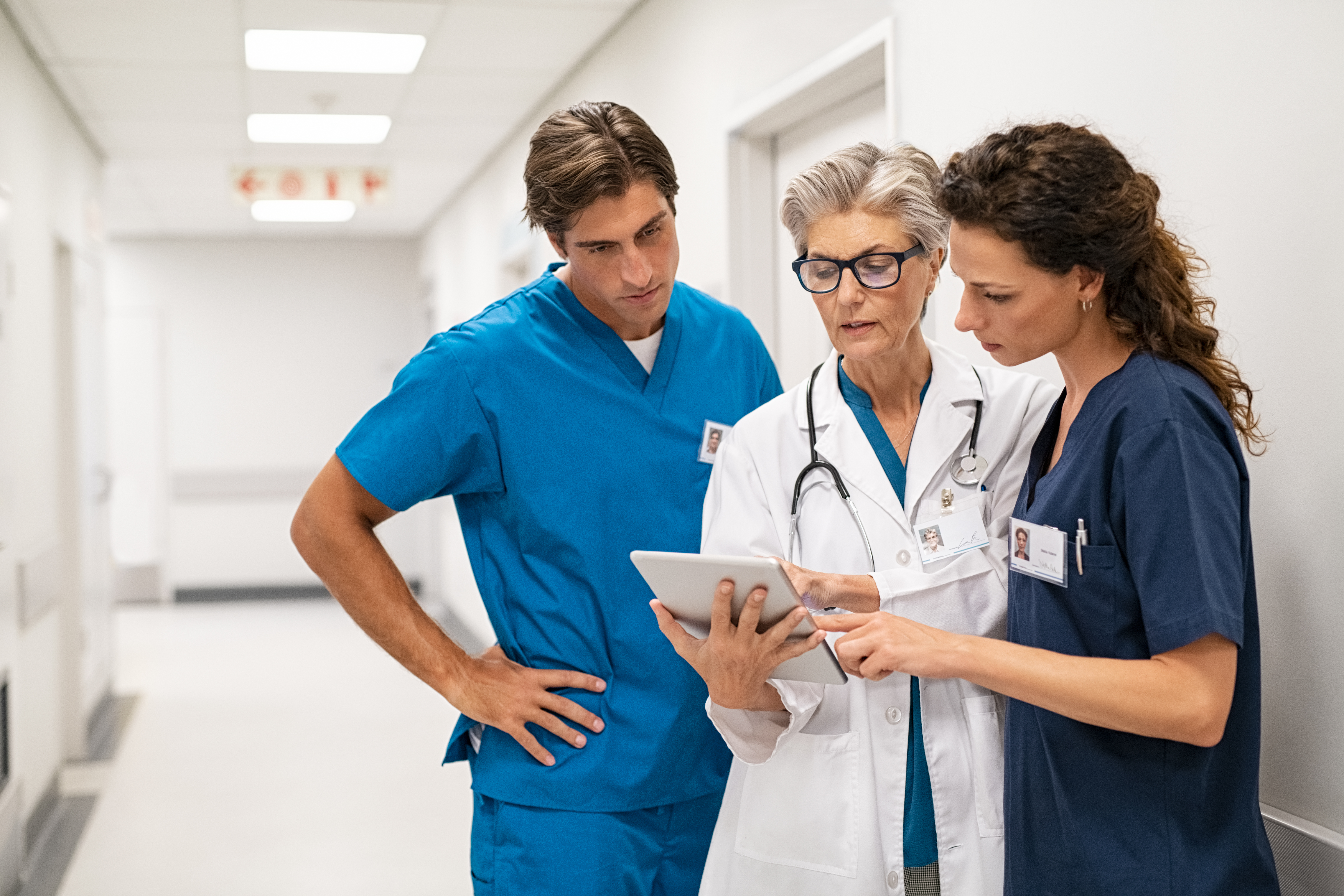 Three healthcare professionals, two in scrubs and one in a white coat, standing in a hospital corridor and discussing information on a tablet.