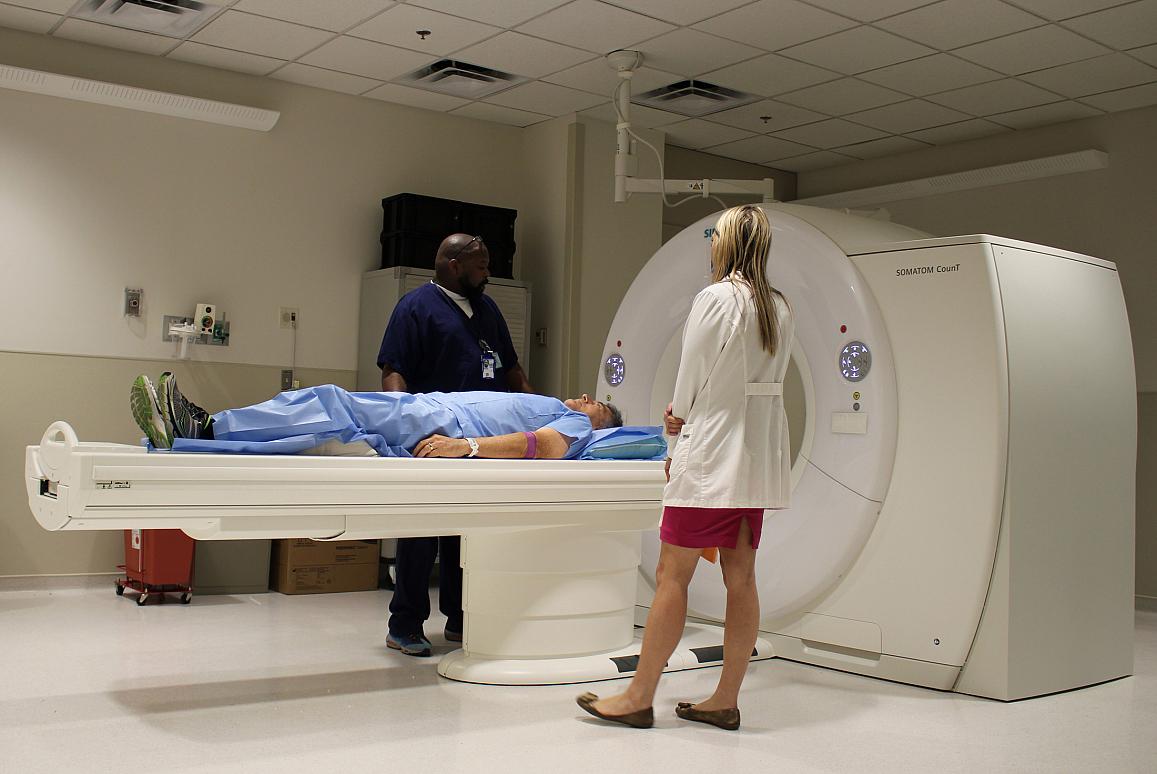 A patient lying on a CT scanner bed with two medical professionals standing nearby, preparing for the scan.