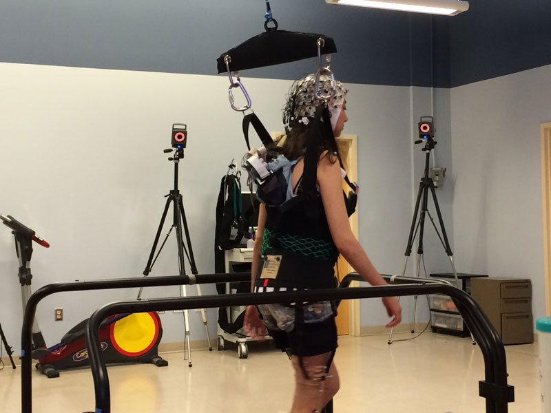 A person in a harness walking on a treadmill in a rehabilitation center, with monitoring equipment and cameras set up around.