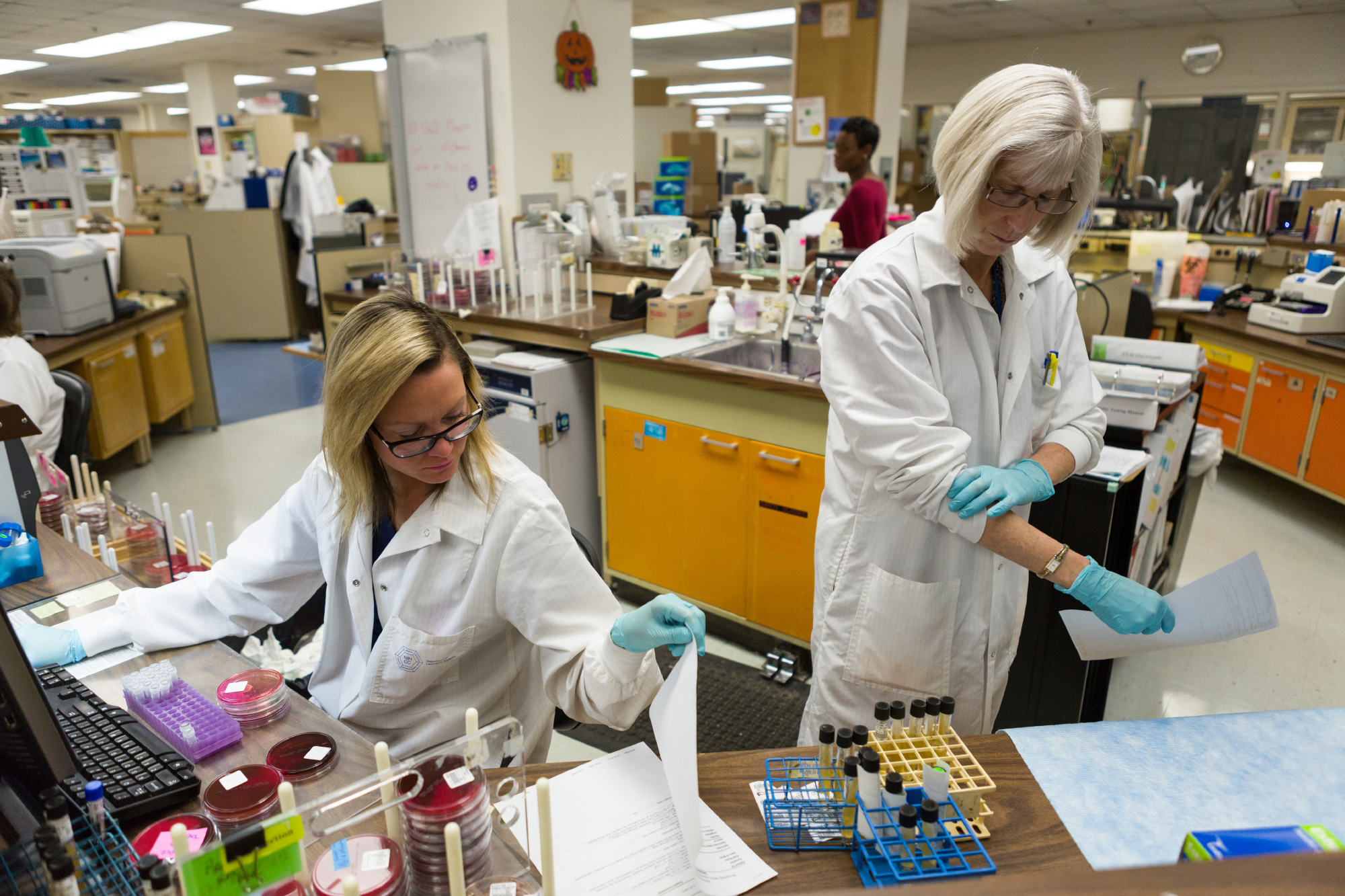 Two laboratory technicians wearing lab coats and gloves work at a desk in a busy lab, one handling paperwork and the other preparing test tubes for analysis.