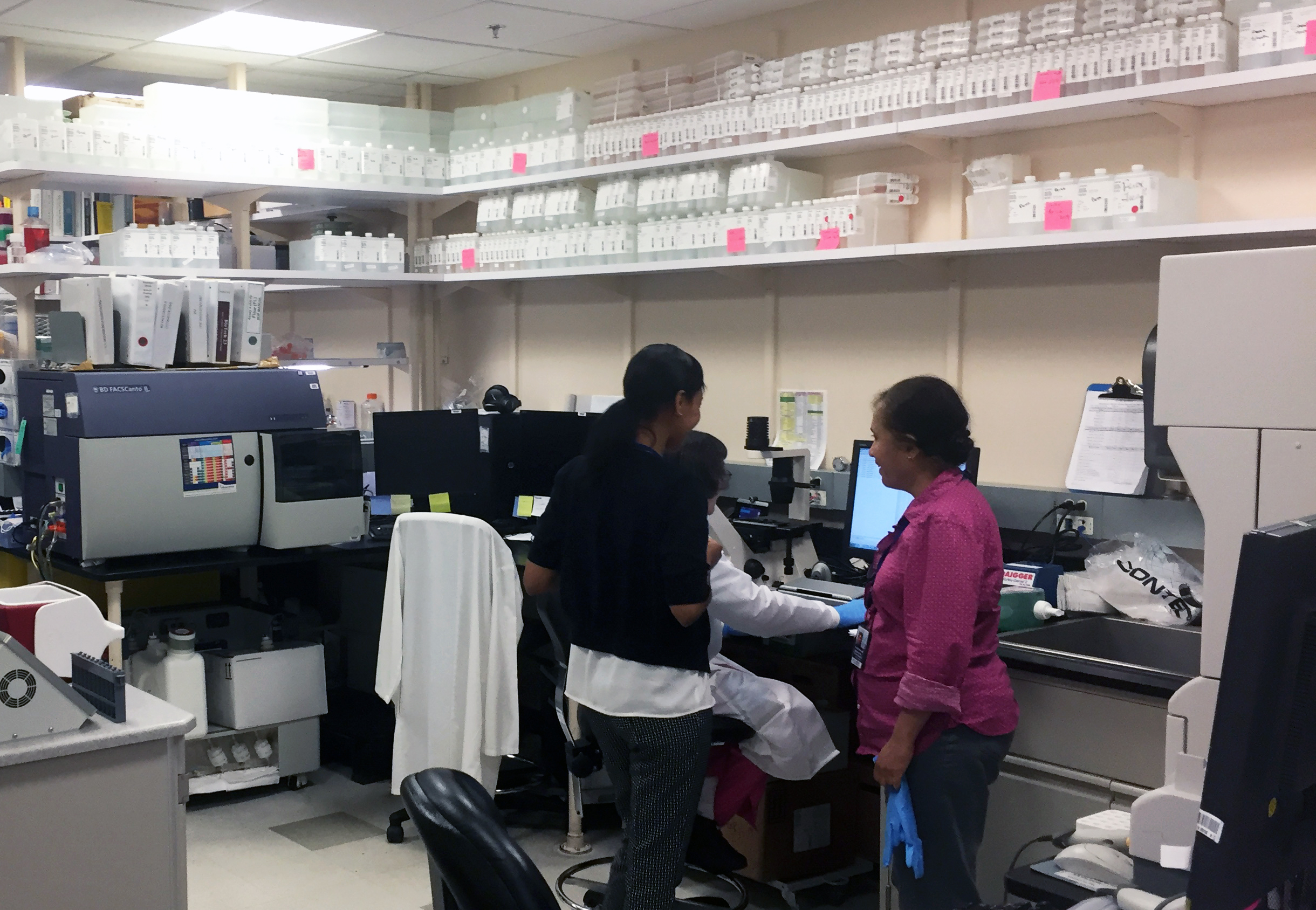 Two laboratory staff members stand beside a colleague seated at a computer in a busy lab filled with equipment and shelves of supplies.