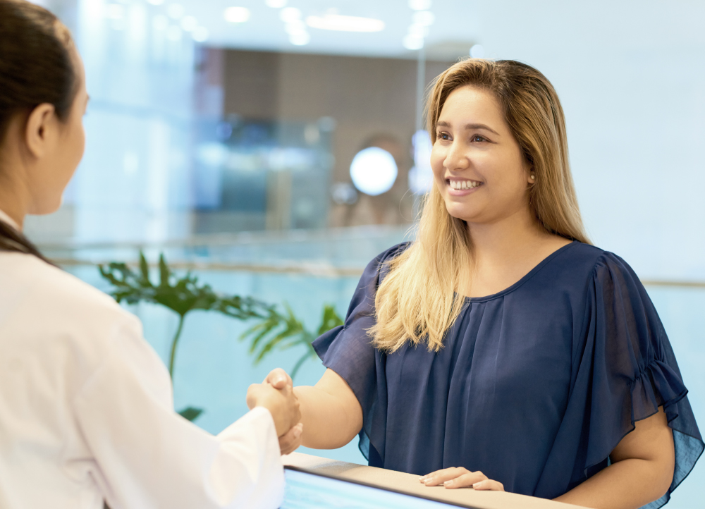 A smiling woman in a blue blouse shakes hands with a healthcare professional across a reception desk in a modern, well-lit office.