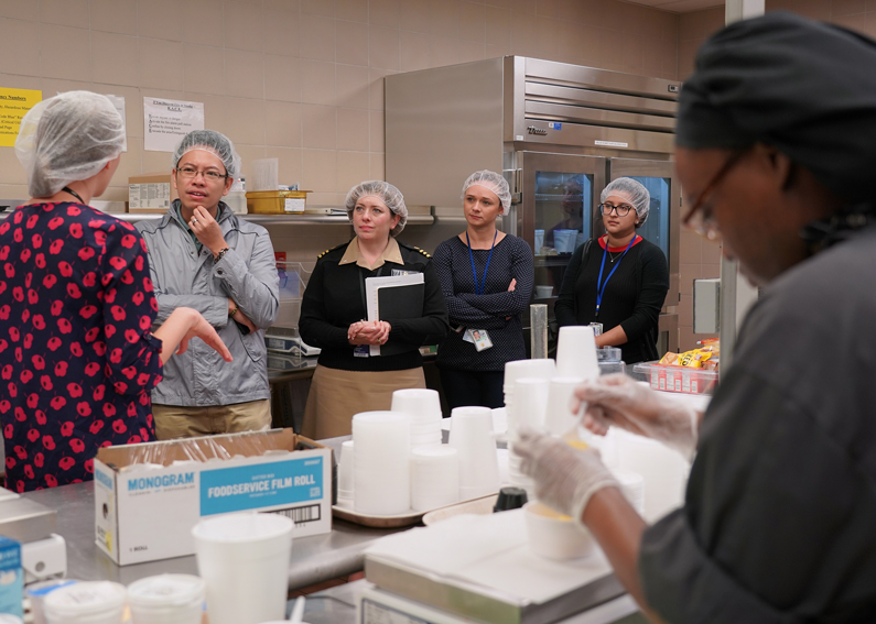 Four people listen to a dietician speak about the Nutrition Department while a employee preps food