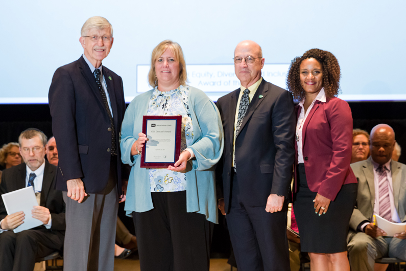 Dr. Francis Collins, Tricia Coffey, Dr. Jim Gilman and Treava Hopkins-Laboy stand on stage as Coffey holds a plaque