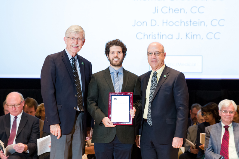 Dr. Francis Collins, Dr. Thomas Bulea and Dr. Jim Gilman stand on stage as Bulea holds a plaque