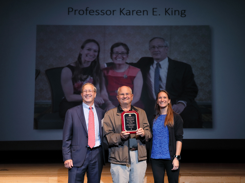 Porter Siems, Paul Ness, Lily SIems on stage. Screen behind has a picture of the Karen King