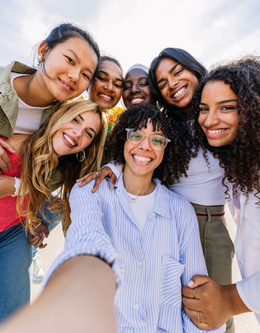 A group of young women smiling and leaning close together while taking a selfie outdoors on a sunny day