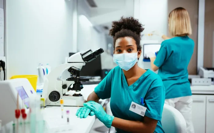 A female lab technician in scrubs and a face mask working at a microscope, with another technician working in the background.