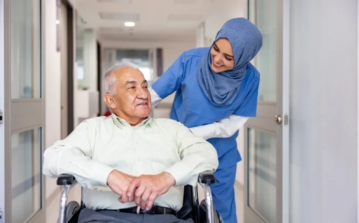 A nurse in scrubs and a hijab smiling at an elderly man in a wheelchair in a hospital corridor.