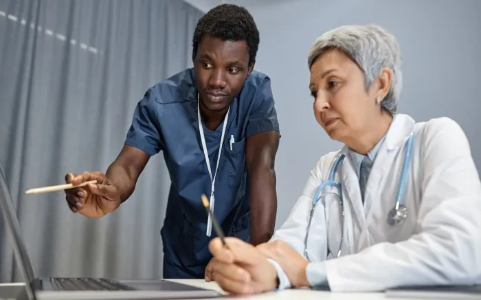 Two healthcare professionals, one in scrubs and the other in a white coat, discussing information on a laptop.