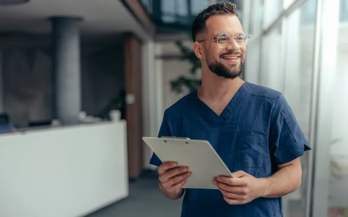 A smiling male healthcare professional in scrubs holding a clipboard in a modern medical office.