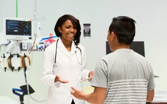 A female doctor in a white coat with a stethoscope speaking with a patient in a medical examination room.