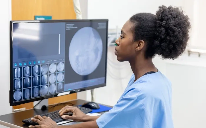 A radiology technician in scrubs working at a computer with medical imaging displayed on the monitor.