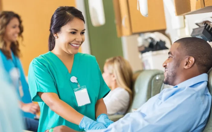 A smiling nurse in scrubs assists a man who is donating blood, while two other people are seen in the background.