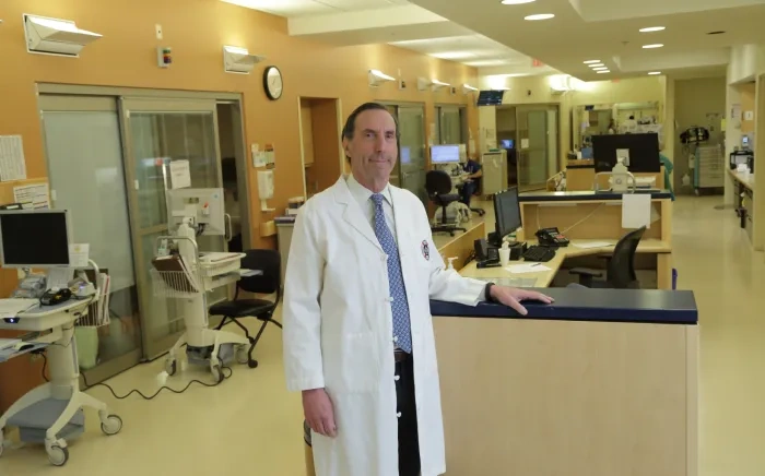 A doctor in a white coat standing in a hospital ward with medical equipment in the background.