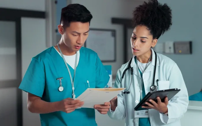 A male nurse in scrubs and a female doctor in a white coat reviewing documents together in a hospital setting.