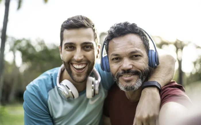 wo smiling men, each wearing headphones around their necks, taking a selfie outdoors with one arm around the other's shoulder.