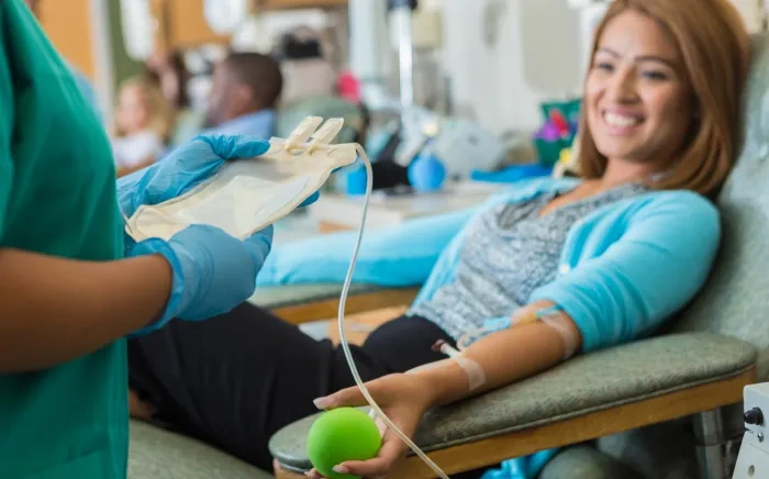 A woman smiling while donating blood, holding a green stress ball, with a medical professional in gloves handling the blood donation bag.