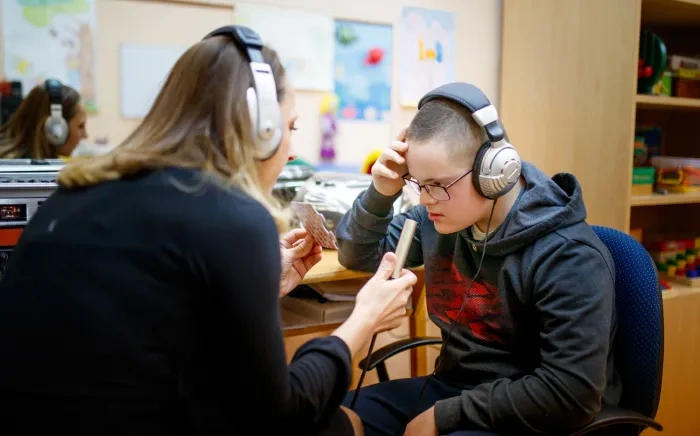 A young person with Down syndrome wearing headphones, participating in a listening activity with a teacher or therapist in a classroom setting.