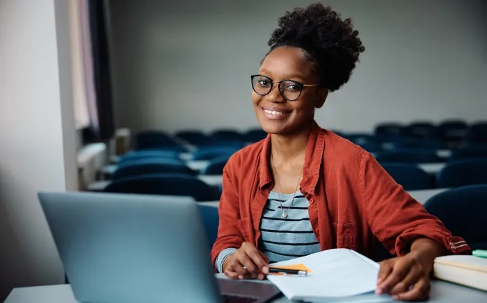 A smiling woman wearing glasses and a red jacket, sitting at a desk with a laptop and holding a pen in a classroom setting.