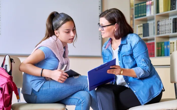 A teacher and a student sitting in a library, engaged in a discussion over an open notebook.