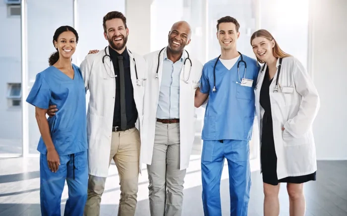 A group of smiling healthcare professionals, including doctors and nurses, stands together in a well-lit hallway, dressed in scrubs and lab coats.