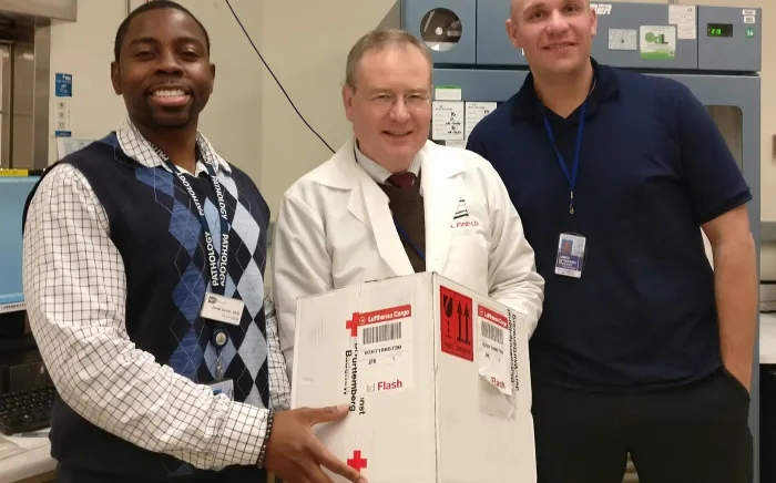 Three smiling healthcare professionals stand together, with one holding a labeled medical package in a clinical or laboratory setting.