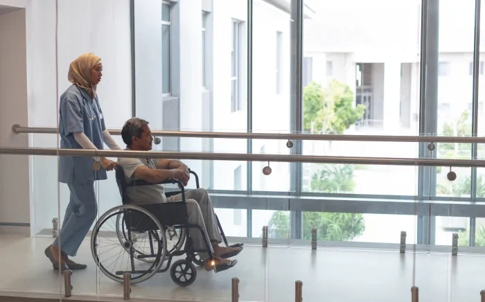 A healthcare worker pushes a patient in a wheelchair down a bright hallway with large windows in a hospital or medical facility.