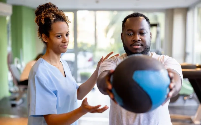 A healthcare professional guides a patient during physical therapy as he holds a medicine ball in a gym-like setting.