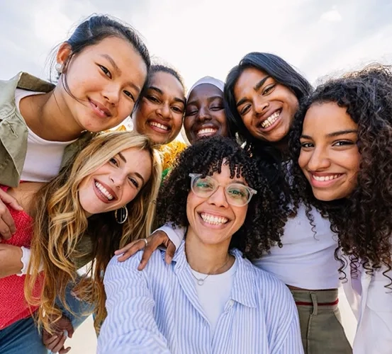 A group of young women smiling and leaning close together while taking a selfie outdoors on a sunny day